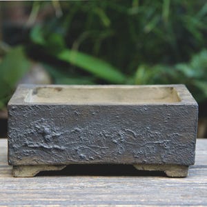 May include: A rectangular, dark gray planter with a textured surface. The planter has a shallow, open top and sits on four small feet. The background is blurred, showing green foliage.