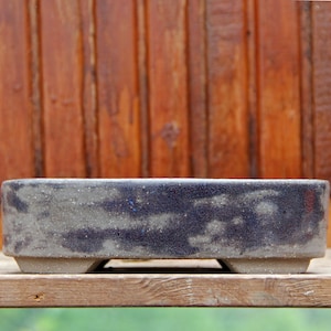 Può includere: Un vaso rettangolare per bonsai con una glassa maculata blu scuro e grigia. Il vaso in ceramica ha un profilo basso e poggia su tre piedini. Lo sfondo è una parete di legno.
