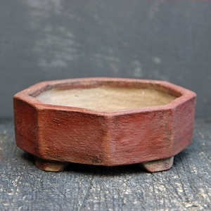 Op de afbeelding: Een rode achthoekige bonsai pot van keramiek met drie kleine voetjes. De pot is leeg en klaar om te planten.