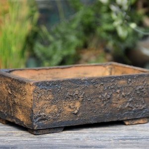 Pode incluir: Um vaso retangular, texturizado, castanho e preto com aparência envelhecida. O vaso tem um perfil baixo e assenta em quatro pés pequenos. O interior é de cor castanha clara. Ideal para pequenas plantas ou suculentas.