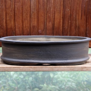 Op de afbeelding: Een lage, ovale bonsai-pot van keramiek met een matte donkergrijze afwerking. De pot heeft een gladde, licht gebogen rand en een brede, ondiepe kom met een getextureerd buitenoppervlak, geschikt voor het planten van kleine bomen.