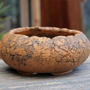 May include: A small, round, brown ceramic planter with a textured surface and a scalloped rim. The planter has a rustic appearance and sits on three small feet. Ideal for succulents or small plants.