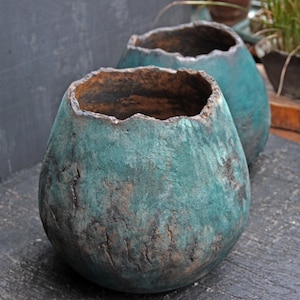 May include: Two teal-colored, handcrafted ceramic pots with a textured, weathered appearance. The pots have a rough, uneven rim and a rounded, bulbous shape. The exterior shows a mix of teal and brown tones, suggesting an aged finish.