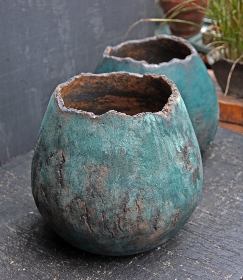 May include: Two teal-colored, handcrafted ceramic pots with a textured, weathered appearance. The pots have a rough, uneven rim and a rounded, bulbous shape. The exterior shows a mix of teal and brown tones, suggesting an aged finish.