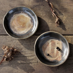 Op de afbeelding: Twee handgemaakte keramische kommen met een rustiek, organisch ontwerp. De kommen hebben een donkere, getextureerde rand en een lichter, gevlekt interieur met tinten bruin en beige. De kommen worden gepresenteerd op een houten oppervlak met gedroogde plantenelementen.