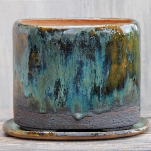 May include: A cylindrical ceramic planter with a matching saucer, featuring a reactive glaze in shades of deep teal, forest green, and earthy brown. The base has a dark, textured, matte finish, while the upper portion displays a glossy drip effect.