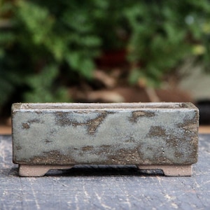 Pode incluir: Um vaso retangular de cerâmica para bonsai com um esmalte verde claro e marrom. O vaso tem uma superfície texturizada e três pequenos pés.