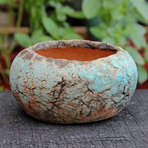 Op de afbeelding: Een kleine, ronde, handgemaakte keramische pot met een getextureerd oppervlak. De pot heeft een mix van turquoise, bruin en zwart, met een oranje binnenkant. Geschikt voor kleine planten of decoratieve doeleinden.