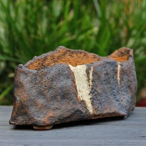 Può includere: Una fioriera rettangolare in ceramica fatta a mano con una superficie ruvida e strutturata. La fioriera presenta un esterno marrone scuro e grigio con un interno marrone più chiaro e una striscia verticale color crema. La fioriera è progettata con un piccolo piede.