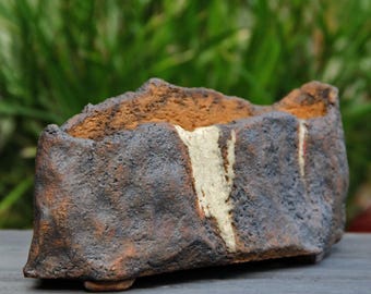 Può includere: Una fioriera rettangolare in ceramica fatta a mano con una superficie ruvida e strutturata. La fioriera presenta un esterno marrone scuro e grigio con un interno marrone più chiaro e una striscia verticale color crema. La fioriera è progettata con un piccolo piede.
