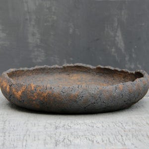 Può includere: Una ciotola in ceramica ovale fatta a mano con una superficie ruvida e strutturata. La ciotola presenta un esterno marrone scuro con accenti marrone chiaro e un bordo leggermente irregolare. L'interno è di colore marrone caldo.