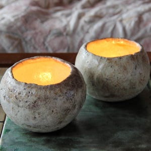 Peut inclure: Deux bougeoirs sphériques texturés en forme de pierre avec de la cire orange-jaune incandescente à l'intérieur. Les bougeoirs sont posés sur une surface verte et bleue, avec un tissu à motifs en arrière-plan. Les bougeoirs ont une esthétique rustique et naturelle.