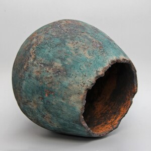 May include: A handcrafted, teal-colored ceramic vessel with a rough, textured surface. The vessel has a large opening on one side, revealing an orange interior. The exterior shows hints of brown and rust.
