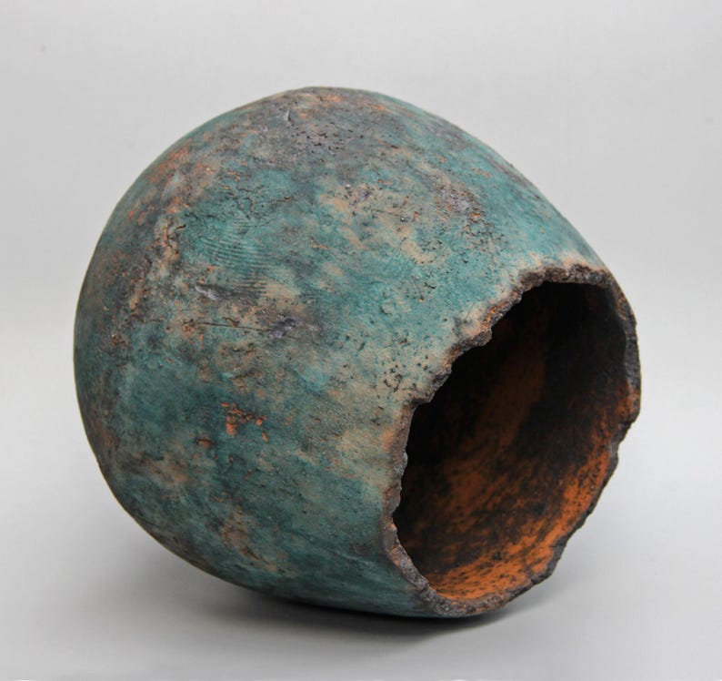 May include: A handcrafted, teal-colored ceramic vessel with a rough, textured surface. The vessel has a large opening on one side, revealing an orange interior. The exterior shows hints of brown and rust.