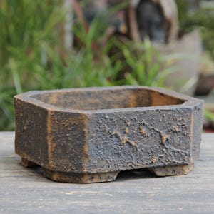 May include: A square, dark brown planter with a textured surface and small feet. The planter has a geometric design and is made of a material that appears to be stone or concrete. The background is blurred, with green foliage.