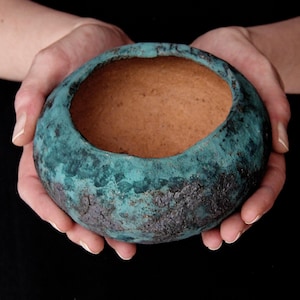 Könnte beinhalten: Eine handgefertigte, runde Keramikschale mit einer strukturierten, türkisfarbenen Außenseite und einem braunen Innenraum. Die Schale wird in zwei Händen vor einem schwarzen Hintergrund gehalten. Die Außenseite der Schale hat ein gealtertes Aussehen.