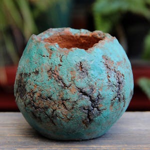 Puede incluir: Una pequeña maceta redonda de cerámica turquesa con superficie texturizada. La maceta tiene un aspecto rugoso y agrietado con detalles en marrón y negro. El interior es de color marrón rojizo cálido. Adecuado para plantas pequeñas o como acento decorativo único.