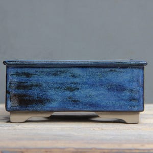 May include: A rectangular, blue ceramic bonsai pot with a glossy finish. The pot has a dark blue and black mottled pattern and sits on four small, light-colored feet. The pot's edges are slightly raised, and the overall design is simple and elegant.