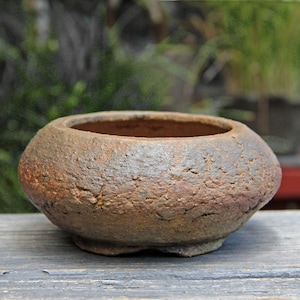 Könnte beinhalten: Kleiner, runder, unglasierter Keramik-Bonsai-Topf mit einer strukturierten, erdfarbenen braunen Oberfläche. Der Topf hat eine breite Öffnung und steht auf drei kleinen Füßen. Das Äußere hat ein raues, natürliches Aussehen, geeignet für Miniaturpflanzen.