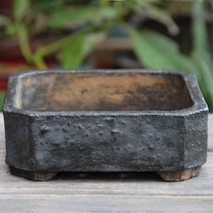 Puede incluir: Una maceta cuadrada de bonsai de cerámica negra con una superficie texturizada rugosa y cuatro pequeños pies. La maceta tiene un borde ligeramente elevado y está hecha de arcilla sin esmaltar.