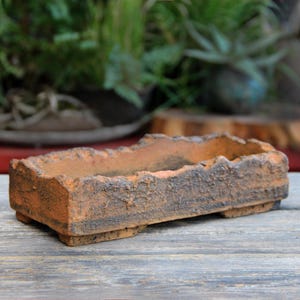 Può includere: Una fioriera rettangolare in terracotta invecchiata con una superficie ruvida e strutturata. La fioriera ha una combinazione di colori marrone scuro e arancione e poggia su piccoli piedini. Ideale per piante grasse o piccole piante.