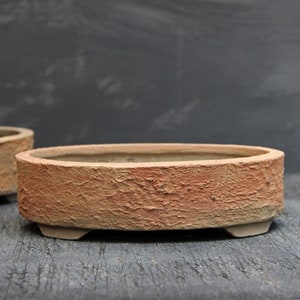 Könnte beinhalten: Ein ovaler Bonsai-Topf aus hellem Braun mit einer rauen, strukturierten Oberfläche. Der Topf hat drei kleine Füße an der Unterseite.