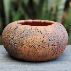 May include: A round, low-profile ceramic bowl with a textured, crackled surface in shades of earthy brown and burnt orange. The vessel features a wide, open top with a thick, uneven rim and a dark, weathered finish across its exterior body.