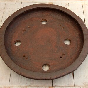 May include: A round, terracotta-colored clay saucer with a raised rim and six evenly spaced holes. The saucer has a rustic, earthy appearance.