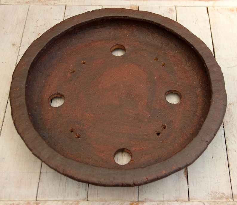 May include: A round, terracotta-colored clay saucer with a raised rim and six evenly spaced holes. The saucer has a rustic, earthy appearance.