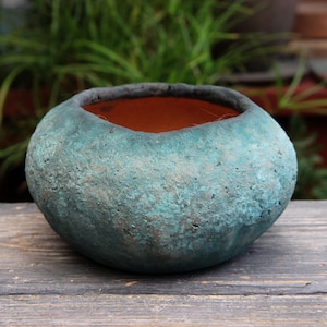 May include: A round, turquoise-colored ceramic planter with a textured surface. The interior is a warm terracotta color, and the rim is dark gray. The planter sits on a weathered wooden surface, with greenery in the background.