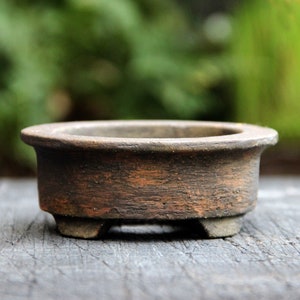 May include: A small, round, dark brown ceramic bonsai pot with a textured, weathered surface. The vessel features a wide, flat rim, a shallow basin, and is supported by three short, sturdy feet at the base, showcasing a rustic, earthy appearance.