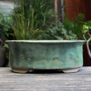 May include: An oval ceramic bonsai pot with a matte, mottled green glaze. The vessel features a slightly flared rim, a textured, uneven surface, and is supported by four small, integrated feet at the base, showcasing a natural, earthy finish.