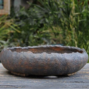 Op de afbeelding: Een ovale keramische plantenbak met een getextureerde, donkerbruine buitenkant en een ruwe, ongelijke rand. De plantenbak staat op een houten oppervlak, met een vage groene achtergrond.