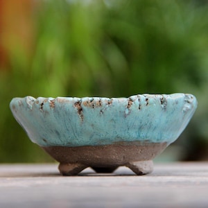 Può includere: Una piccola ciotola in ceramica fatta a mano con una glassa turchese e un bordo strutturato. La ciotola ha una base marrone scuro ed è sostenuta da tre piccoli piedini. Probabilmente viene utilizzata per piante o piccoli oggetti decorativi.