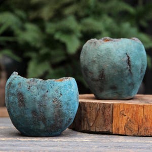 Op de afbeelding: Twee kleine, ronde, blauwgroene plantenbakken met een ruw, verweerd en getextureerd oppervlak. De plantenbakken hebben een onregelmatige, organische rand en een donkere, gevlekte afwerking die doet denken aan steen, waarbij de een iets achter de ander staat.