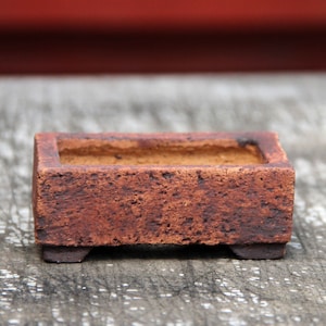 May include: A small, rectangular, brown ceramic planter with a textured surface. The planter has a low profile and sits on four small feet. The interior is a lighter brown color. Suitable for small plants or succulents.