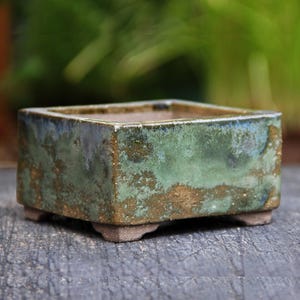 Puede incluir: Una maceta cuadrada de bonsái de cerámica con un esmalte verde y dorado. La maceta tiene cuatro pequeñas patas en la parte inferior.