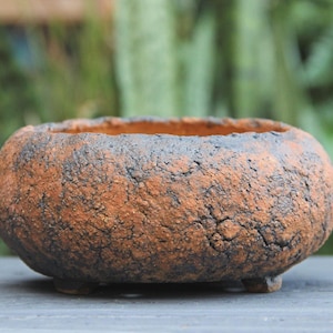 May include: A round, low-profile ceramic planter with a rough, textured surface in shades of burnt orange and charcoal black. The vessel features a wide, open top and rests on three small, integrated feet, showcasing a weathered, earthy appearance.