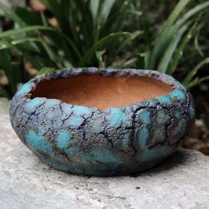 May include: A handcrafted ceramic bowl with a textured, turquoise and dark brown exterior. The bowl has a rough, cracked surface and a smooth, light brown interior. Suitable for plants or decorative purposes.