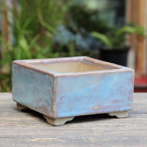 Może przedstawiać: Prostokątna ceramiczna donica z jasnoniebieską glazurą i brązową obwódką. Donica ma kwadratowy kształt i stoi na czterech małych nóżkach. Tło jest rozmyte, z widocznymi zielonymi liśćmi.