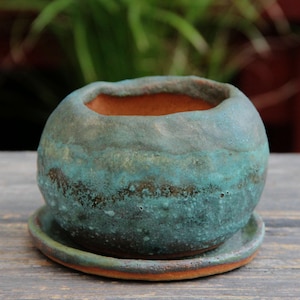 May include: A small, round ceramic planter with a matching saucer. The planter has a textured, turquoise and brown glaze. The interior is a warm terracotta color. The planter is designed for small plants and succulents.