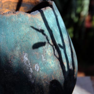 May include: A weathered, teal-colored ceramic pot with an orange rim. The pot's surface has a textured appearance with white patches. Shadows of branches are cast across the pot's surface, creating a contrast of light and dark.