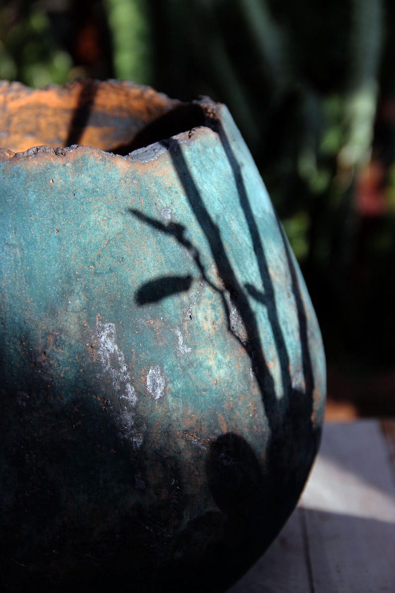 May include: A weathered, teal-colored ceramic pot with an orange rim. The pot's surface has a textured appearance with white patches. Shadows of branches are cast across the pot's surface, creating a contrast of light and dark.