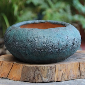 May include: A teal-colored, textured ceramic planter with a wide opening. The planter has a rough, stone-like appearance and sits on a wooden base. The interior of the planter is a warm, earthy brown color. Ideal for succulents or small plants.