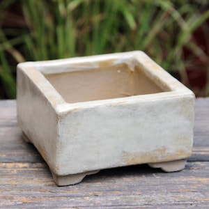 Può includere: Un vaso quadrato in ceramica bianco sporco dall'aspetto invecchiato. Il vaso ha un'apertura rettangolare e poggia su quattro piedini. Il colore neutro e il design semplice lo rendono adatto a varie piante e stili di arredamento.