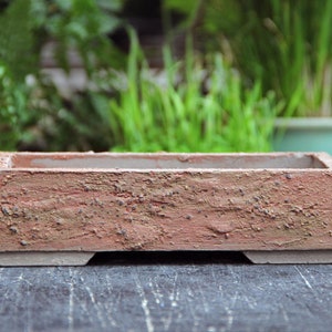 Puede incluir: Una maceta rectangular de bonsai de color terracota con una superficie rugosa y texturizada. La maceta tiene un borde ligeramente elevado y tres pequeños pies en la parte inferior.