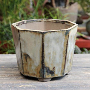 May include: A hexagonal ceramic planter featuring a reactive glaze with mottled shades of cream, beige, and dark brown. The pot has a wide, open top, defined vertical edges, and rests on small, integrated feet at the base, showcasing a smooth finish.
