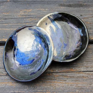 Peut inclure: Deux bols en céramique faits à la main avec une finition brillante. Les bols présentent un mélange de couleurs bleu, gris et noir, créant un effet marbré. Les bols sont posés sur une surface en bois vieilli.