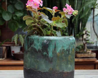 Può includere: Un vaso cilindrico in ceramica con smalto reattivo caratterizzato da una sfumatura di turchese e verde acqua che cola su una base scura e testurizzata color carbone. Il vaso contiene una piccola pianta con foglie verdi e fiori rosa.
