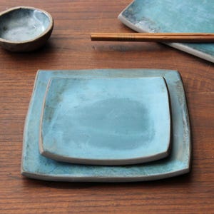 Puede incluir: Dos platos cuadrados de cerámica, uno encajado dentro del otro, ambos con un esmalte azul claro. Los platos tienen un borde ligeramente redondeado y una textura sutil y desigual.